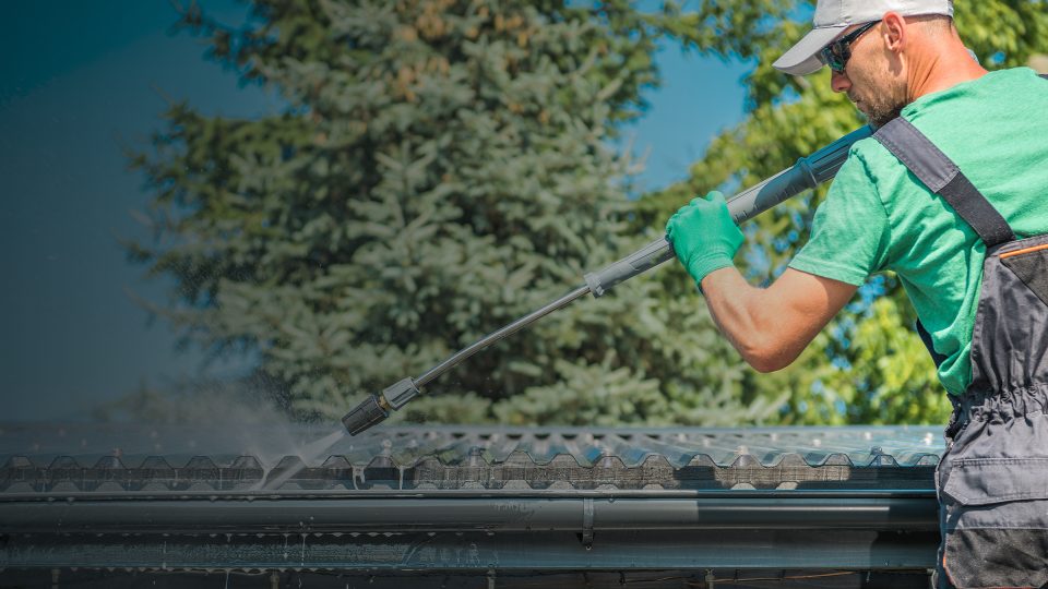 A technician wearing work overalls, gloves, and sunglasses using a pressure washer wand to spray down a corrugated roof panel and clean out the gutter underneath.