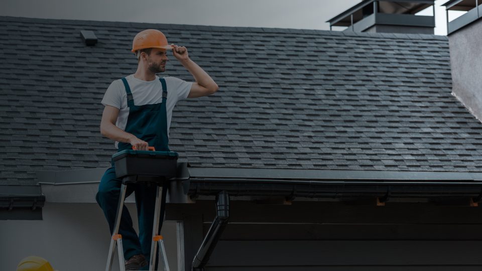 A professional roofer in a hard hat and blue work overalls carrying a toolbox while standing at the top of a ladder to inspect dark asphalt roof shingles.